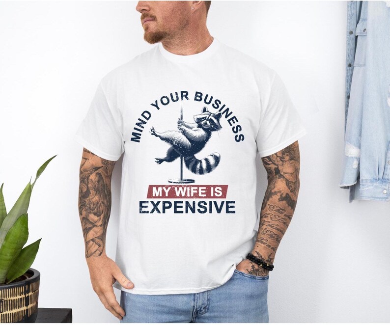 Puede incluir: Camiseta blanca con un mapache en una barra con el texto "MIND YOUR BUSINESS" arriba. Debajo, el texto "MY WIFE IS EXPENSIVE". Es una prenda casual, adecuada para el uso diario.