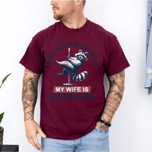 Puede incluir: Camiseta burdeos con un gr&aacute;fico de un mapache bailando en barra. El texto en la camiseta dice "MIND YOUR BUSINESS" encima del mapache y "MY WIFE IS EXPENSIVE" debajo.