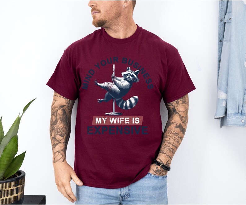 Puede incluir: Camiseta burdeos con un gr&aacute;fico de un mapache bailando en barra. El texto en la camiseta dice "MIND YOUR BUSINESS" encima del mapache y "MY WIFE IS EXPENSIVE" debajo.