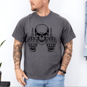 May include: Dark gray t-shirt featuring a black skull graphic with both hands giving the middle finger. The skull has a menacing grin. The shirt is a casual style, suitable for everyday wear.