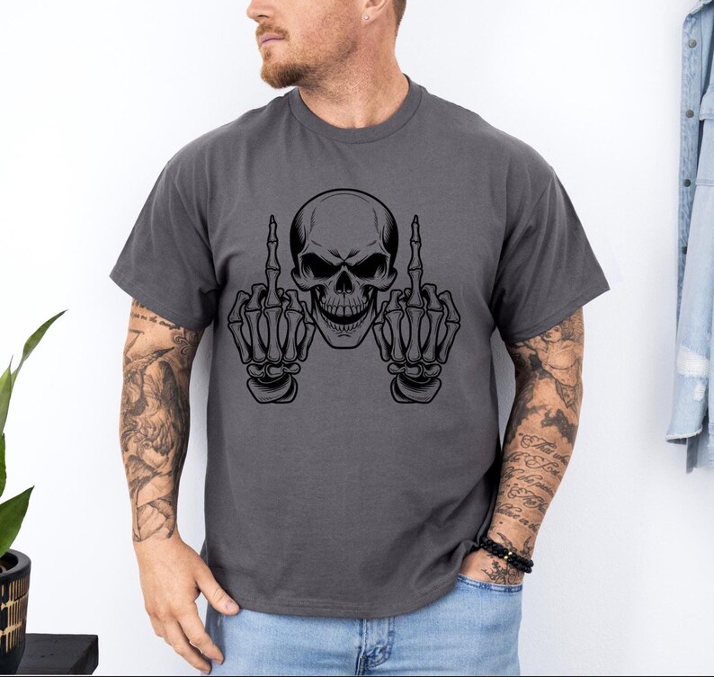 May include: Dark gray t-shirt featuring a black skull graphic with both hands giving the middle finger. The skull has a menacing grin. The shirt is a casual style, suitable for everyday wear.