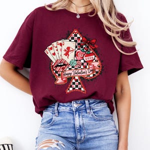May include: Burgundy t-shirt featuring a Valentine's Day design. The graphic includes playing cards, lips, hearts, and the text "Happy Valentine's Day" on a banner. The design is set against a black and white checkered spade shape.