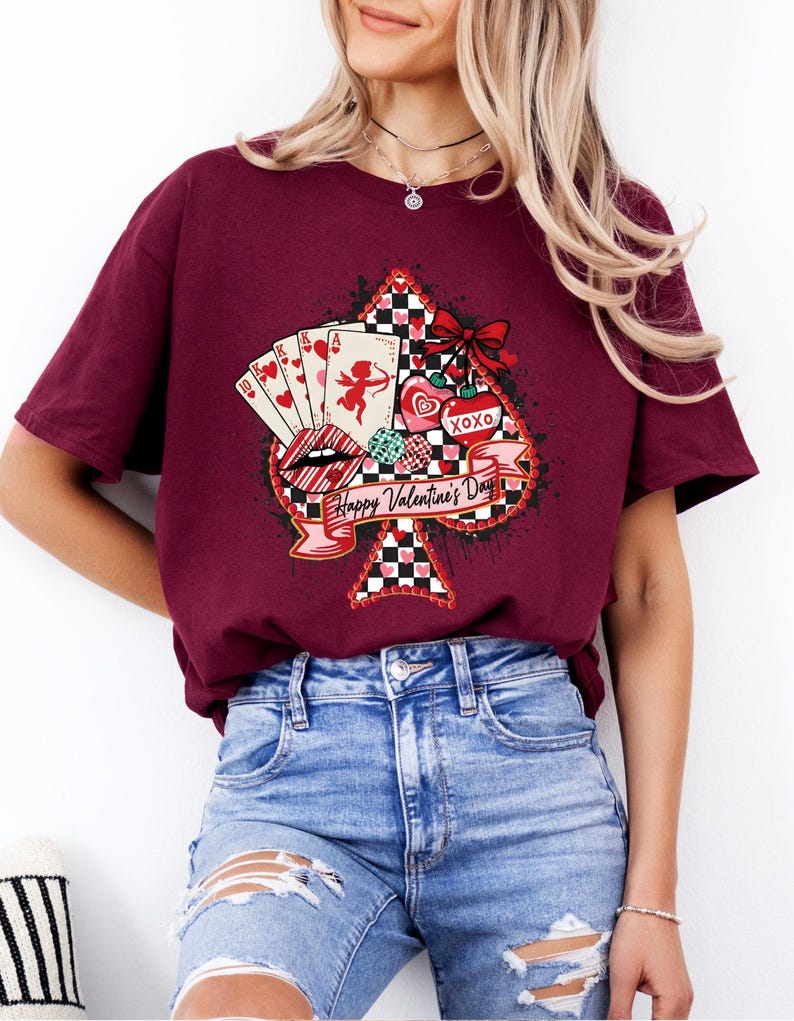 May include: Burgundy t-shirt featuring a Valentine's Day design. The graphic includes playing cards, lips, hearts, and the text "Happy Valentine's Day" on a banner. The design is set against a black and white checkered spade shape.