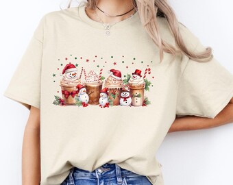 May include: Beige t-shirt featuring a watercolor design of five festive coffee cups, each adorned with whipped cream, a snowman, and a Santa hat. Red and green holiday accents complete the design, perfect for the Christmas season.