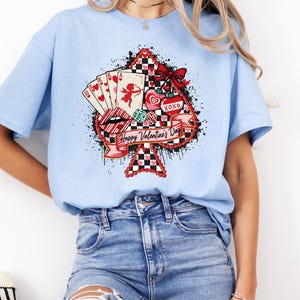 May include: A light blue t-shirt featuring a Valentine's Day design. The design includes a playing card suit shape with a red and black checkered pattern, playing cards, lips, hearts, and the text "Happy Valentine's Day".