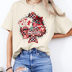 May include: Cream-colored t-shirt featuring a Valentine's Day design. The graphic includes a spade shape with playing cards, a cupid, lips, hearts, and the text "Happy Valentine's Day" and "XOXO". The design has red, black, and white accents.