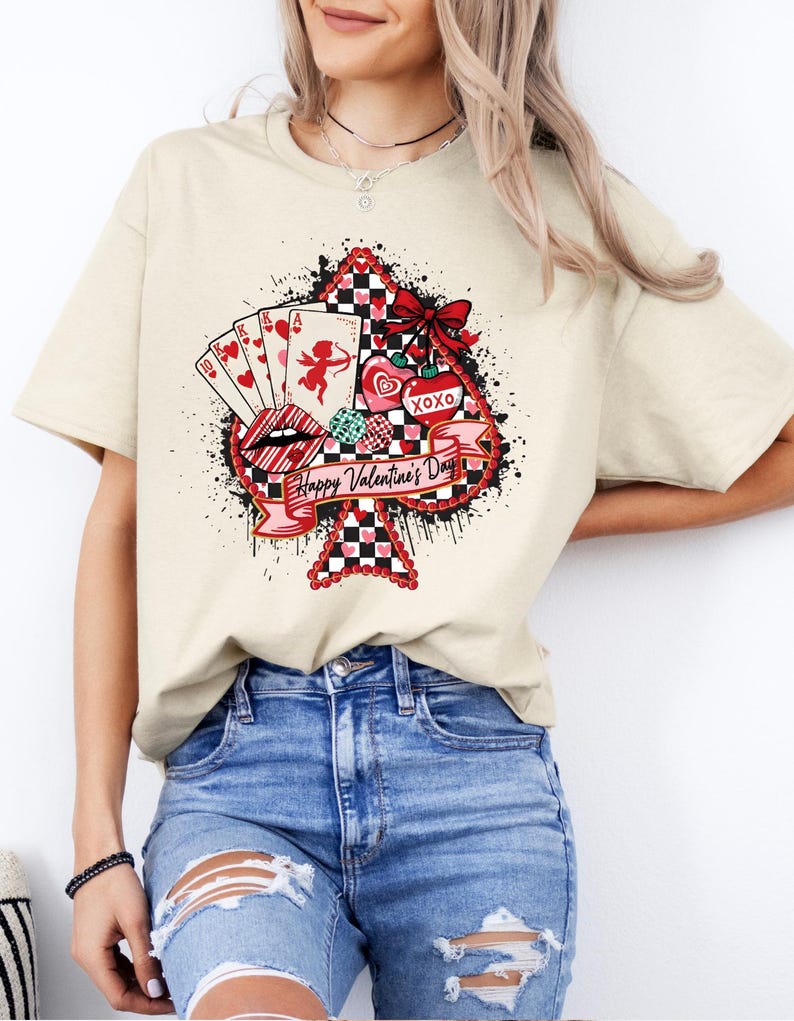 May include: Cream-colored t-shirt featuring a Valentine's Day design. The graphic includes a spade shape with playing cards, a cupid, lips, hearts, and the text "Happy Valentine's Day" and "XOXO". The design has red, black, and white accents.