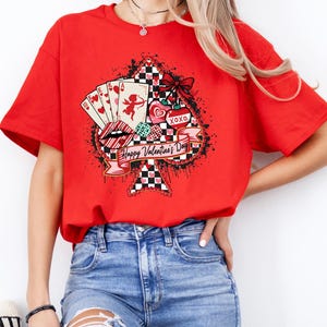 May include: A red t-shirt featuring a graphic design for Valentine's Day. The design includes playing cards, lips, hearts, and the text "Happy Valentine's Day" on a banner. The shirt has short sleeves.