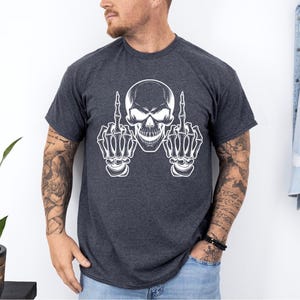 May include: Dark gray t-shirt featuring a white skull graphic with skeletal hands giving the middle finger. The design is centered on the shirt. The shirt has short sleeves and a crew neck.