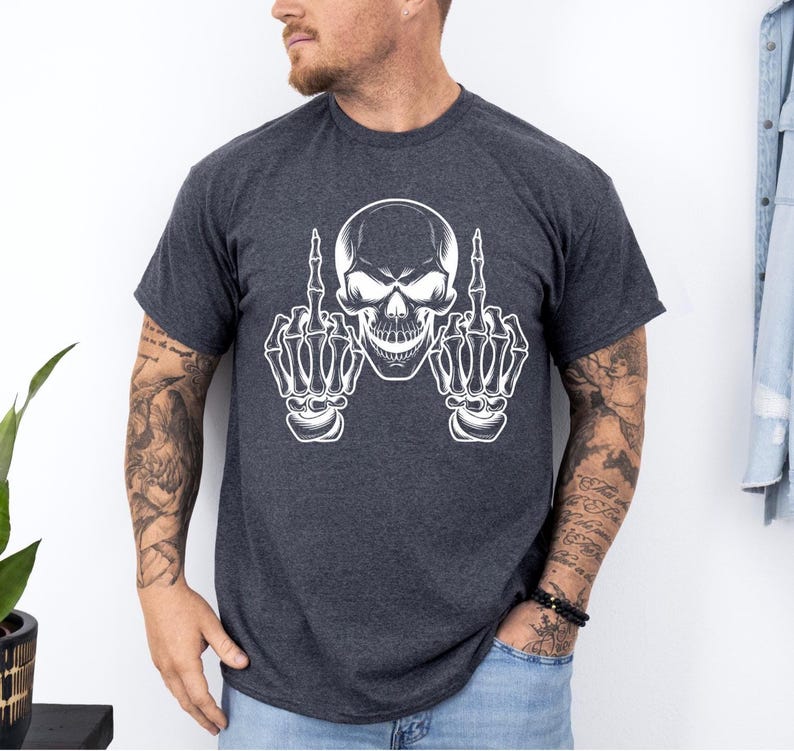 May include: Dark gray t-shirt featuring a white skull graphic with skeletal hands giving the middle finger. The design is centered on the shirt. The shirt has short sleeves and a crew neck.