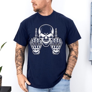 May include: Navy blue t-shirt featuring a white skull graphic with skeletal hands giving the middle finger. The design is centered on the shirt. The shirt has short sleeves and a crew neck.