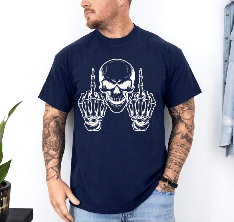 May include: Navy blue t-shirt featuring a white skull graphic with skeletal hands giving the middle finger. The design is centered on the shirt. The shirt has short sleeves and a crew neck.