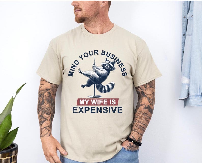 Puede incluir: Camiseta beige con un dise&ntilde;o gr&aacute;fico azul marino y rojo de un mapache haciendo pole dance. El texto dice "MIND YOUR BUSINESS" arriba y "MY WIFE IS EXPENSIVE" abajo.