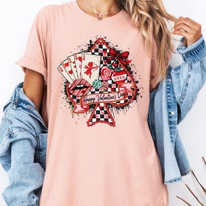 May include: A light pink t-shirt featuring a large ace of spades design with Valentine's Day graphics. The design includes playing cards, hearts, lips, and the text "Happy Valentine's Day". The shirt has rolled-up sleeves.