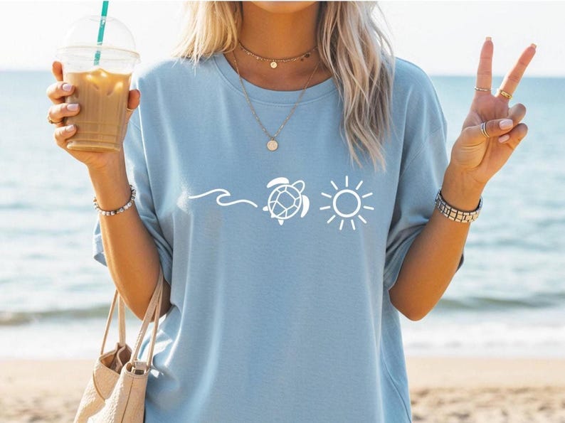 May include: Light blue t-shirt with a white graphic of a wave, a turtle, and a sun. The shirt is made of a soft material. The person is holding a drink in a clear plastic cup with a straw and a beige tote bag.