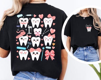 May include: Black t-shirt featuring a whimsical dental-themed design. The back of the shirt showcases a pattern of smiling cartoon teeth, pink hearts, and dental tools. A smaller version of the tooth graphic is on the front. The shirt is paired with blue jeans.