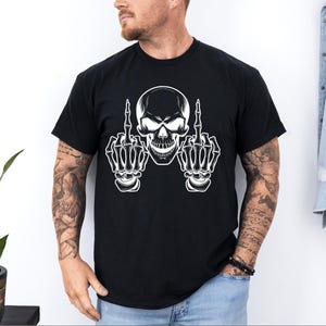 May include: Black t-shirt featuring a white skull graphic with skeletal hands giving the middle finger. The skull has detailed shading and the hands are fully rendered. The shirt is a classic crew neck style.