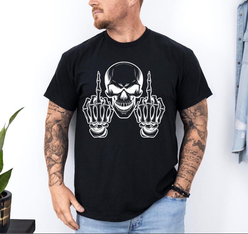 May include: Black t-shirt featuring a white skull graphic with skeletal hands giving the middle finger. The skull has detailed shading and the hands are fully rendered. The shirt is a classic crew neck style.
