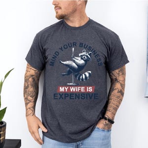 Puede incluir: Camiseta gris oscuro con un dise&ntilde;o de mapache bailando en barra. El texto dice "MIND YOUR BUSINESS" encima del mapache y "MY WIFE IS EXPENSIVE" debajo. La camiseta es de cuello redondo.