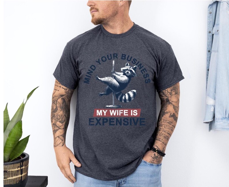 Puede incluir: Camiseta gris oscuro con un dise&ntilde;o de mapache bailando en barra. El texto dice "MIND YOUR BUSINESS" encima del mapache y "MY WIFE IS EXPENSIVE" debajo. La camiseta es de cuello redondo.