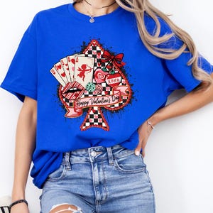 May include: Royal blue t-shirt featuring a Valentine's Day design. The graphic includes a spade shape with playing cards, a cupid, hearts, and the text "Happy Valentine's Day". A red bow and black and white checkered pattern complete the design.