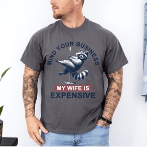 Puede incluir: Camiseta gris oscuro con un gr&aacute;fico de un mapache bailando en barra. El texto dice "MIND YOUR BUSINESS" encima del mapache y "MY WIFE IS EXPENSIVE" debajo. La camiseta es de estilo casual.