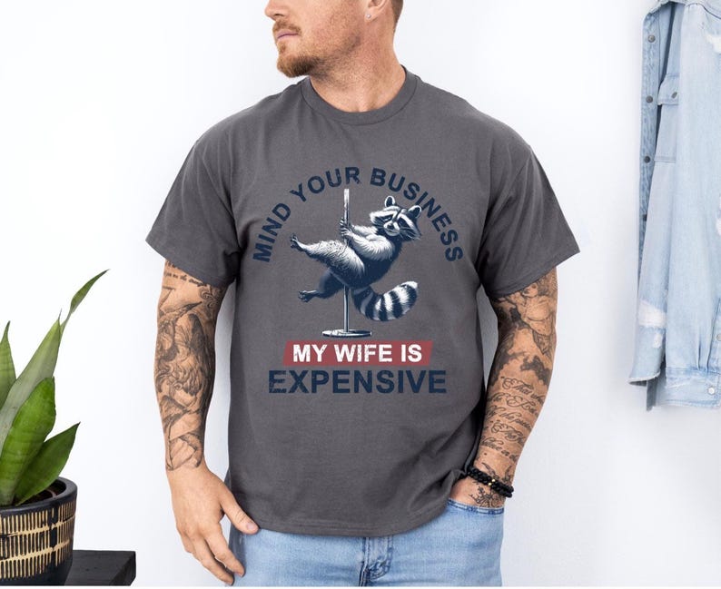 Puede incluir: Camiseta gris oscuro con un gr&aacute;fico de un mapache bailando en barra. El texto dice "MIND YOUR BUSINESS" encima del mapache y "MY WIFE IS EXPENSIVE" debajo. La camiseta es de estilo casual.