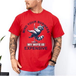 Puede incluir: Camiseta roja con un dise&ntilde;o de mapache en un poste, con el texto "MIND YOUR BUSINESS" y "MY WIFE IS EXPENSIVE" en azul marino. El mapache est&aacute; representado en tonos grises y blancos.