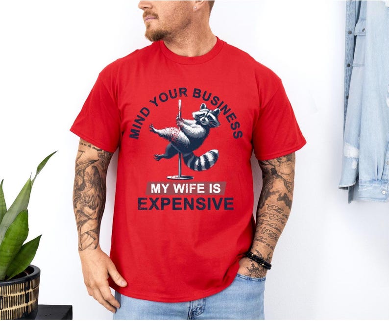 Puede incluir: Camiseta roja con un dise&ntilde;o de mapache en un poste, con el texto "MIND YOUR BUSINESS" y "MY WIFE IS EXPENSIVE" en azul marino. El mapache est&aacute; representado en tonos grises y blancos.