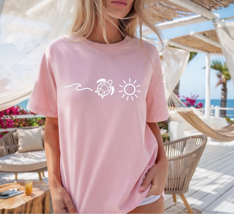 May include: A light pink t-shirt featuring a white graphic of a wave, a sea turtle, and a sun. The shirt has short sleeves and a crew neck. The design is simple and evokes a beach or summer theme.