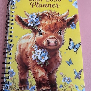 Op de afbeelding: Een spiraalgebonden planner met een gele kaft met een illustratie van een bruin Highland-kalf, blauwe bloemen en vlinders. De tekst 2027-2030 Planner staat bovenaan. Het ontwerp bevat een bloemstuk op de kop en borst van het kalf.