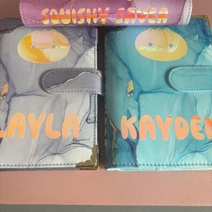 May include: Three rectangular binders with marble-patterned covers. One purple binder at the top reads SQUISHY SAVER. Two binders below feature gold-toned corner accents and magnetic closures, with the names LAYLA and KAYDEN printed in orange.