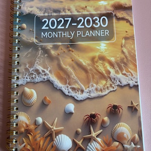 May include: A spiral-bound monthly planner with a cover featuring a sunset beach scene, including ocean waves, seashells, starfish, and small crabs. The text 2027-2030 MONTHLY PLANNER is printed in white within a rectangular frame on the upper portion.