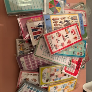 May include: A collection of colourful cards with various designs and prices. The cards feature images of animals, flowers, and other objects. The cards are arranged in a pile on a pink surface.
