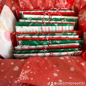 May include: A collection of rectangular envelopes wrapped in red and green Christmas-themed paper with white snowflake patterns. Each envelope is tied with red and white baker's twine and nestled inside a box lined with red tissue paper.