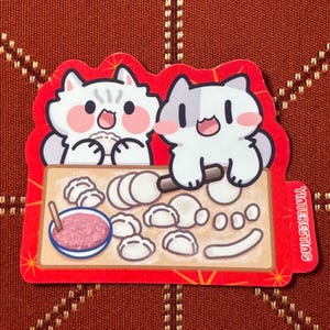 May include: Two white cats with pink cheeks are making dumplings. One cat is holding a rolling pin and the other is holding a dumpling. The cats are on a red background with the text "YUM!" in the corner.