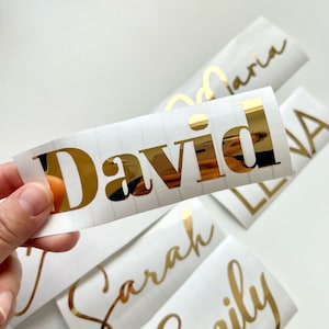 May include: Gold foil name stickers with the names David, Sarah, Emily, and Maria. The stickers are on a white background.