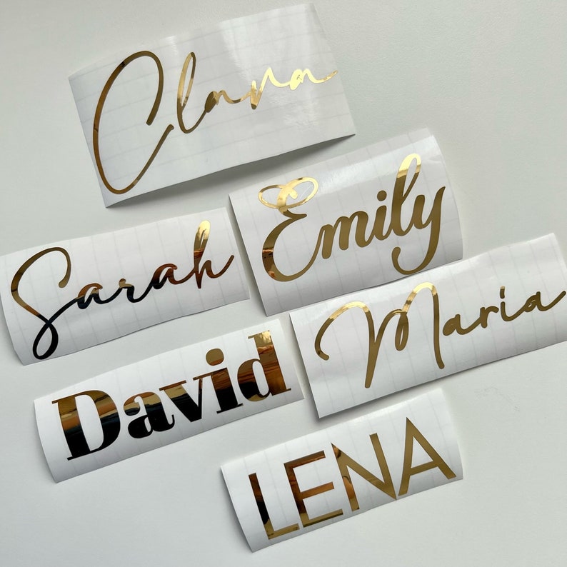 May include: Six white labels with gold lettering. The names on the labels are:  Clara, Emily, Sarah, David, Maria, and Lena.