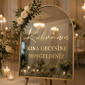 May include: An arched mirror sign with a gold frame, decorated with white roses and green leaves. The text in gold reads: Kübra'nın KINA GECESİNE HOŞGELDİNİZ 09 | 09 | 2024. The sign is displayed on a stand, reflecting a warm, softly lit indoor event space.