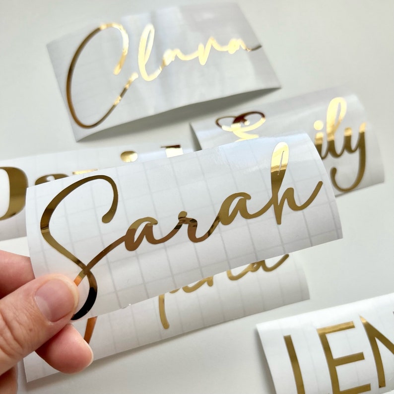 May include: Gold foil personalized name stickers on white sheets. The names are written in a cursive script font. The names are Sarah, Emma, Lily, and Jen.