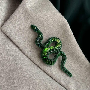May include: A green snake brooch made of beads and rhinestones. The snake is coiled and has a detailed design.