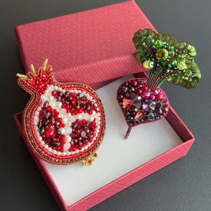 Puede incluir: Dos broches de cuentas en una caja roja. Un broche es una granada roja con detalles blancos y dorados. El otro broche es una remolacha roja con hojas verdes.