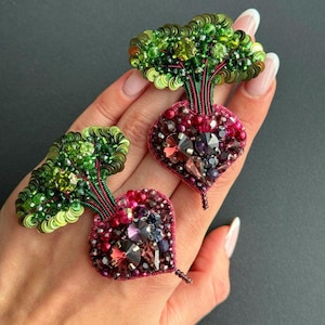 May include: Two handmade beaded brooches in the shape of beets. The beetroots are a deep red-purple color, adorned with various beads and crystals. The green leaves are made of sequins and beads. The brooches are detailed and eye-catching.