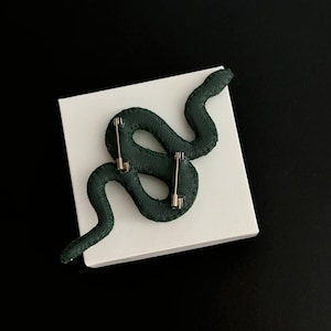 May include: A dark green snake-shaped pin with a silver pin back. The pin is on a white surface.