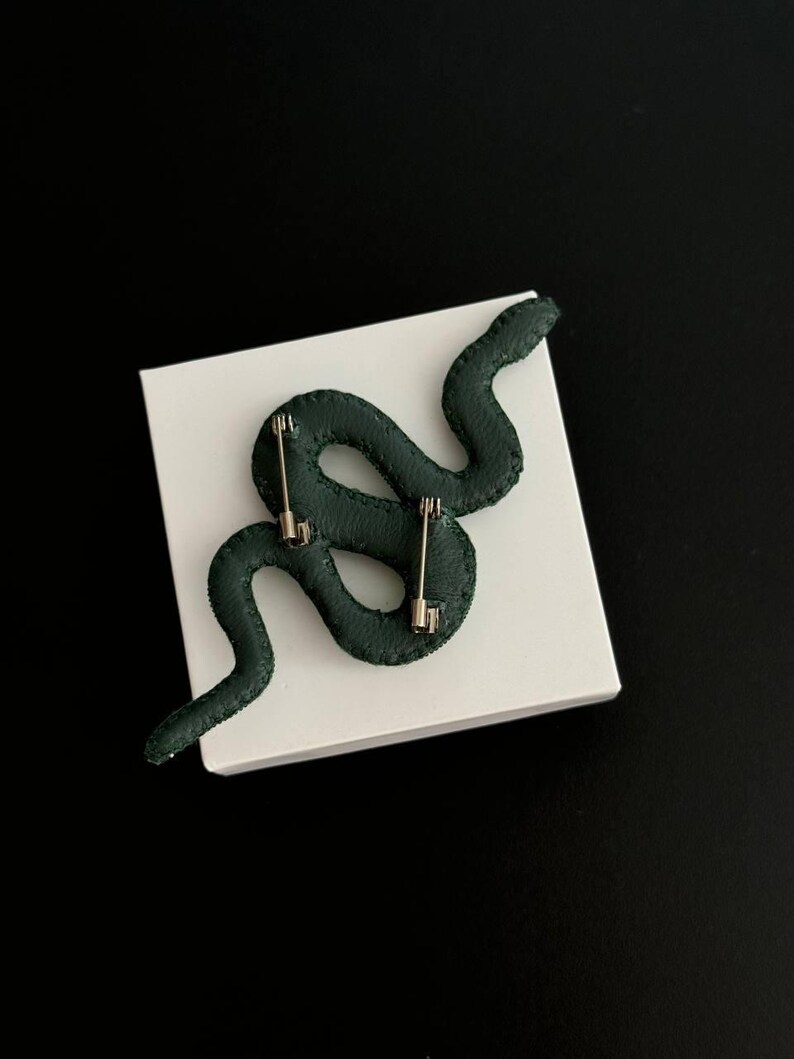 May include: A dark green snake-shaped pin with a silver pin back. The pin is on a white surface.