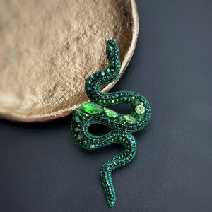 May include: A green snake brooch made of beads and rhinestones. The snake is coiled and has a detailed design.