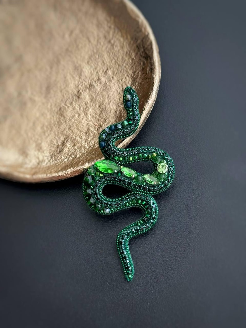 May include: A green snake brooch made of beads and rhinestones. The snake is coiled and has a detailed design.