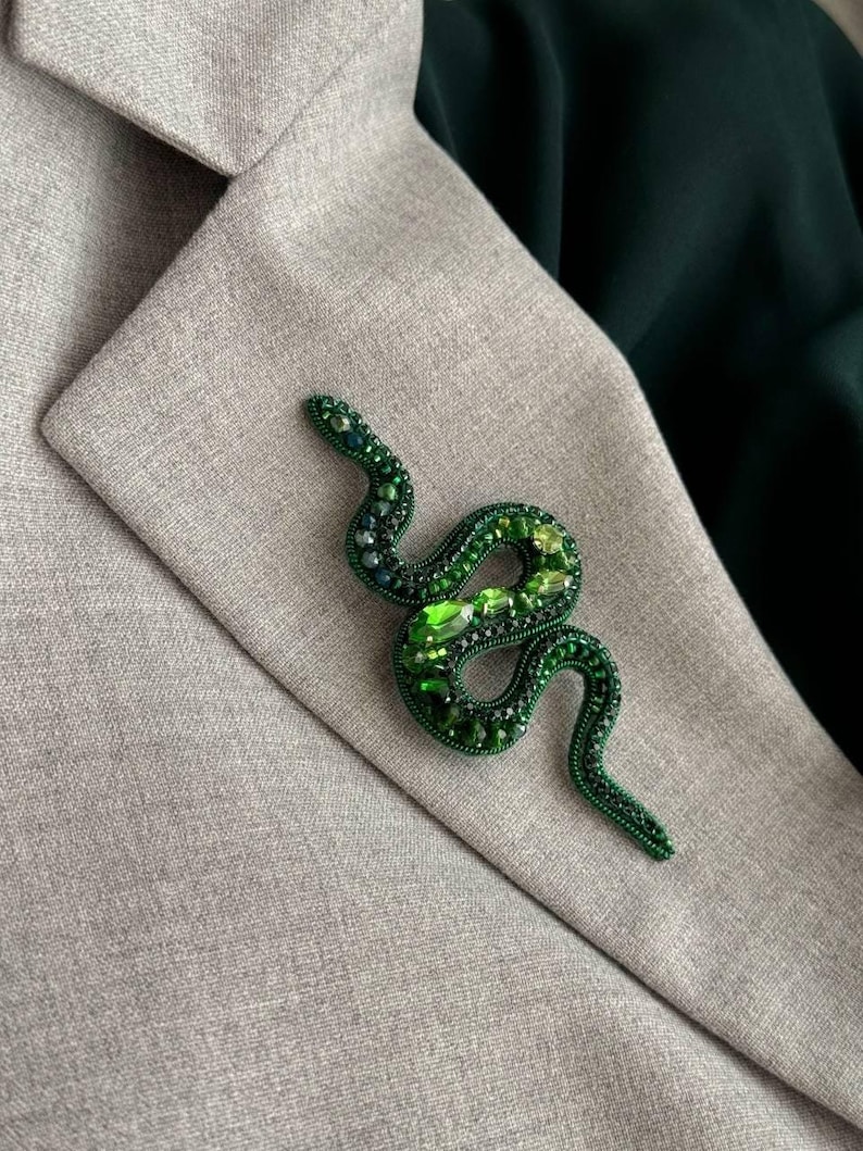 May include: A green snake brooch made of beads and rhinestones. The snake is coiled and has a detailed design.