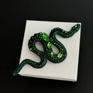 May include: A green snake brooch made of beads and rhinestones. The snake is curled up in a coil shape.