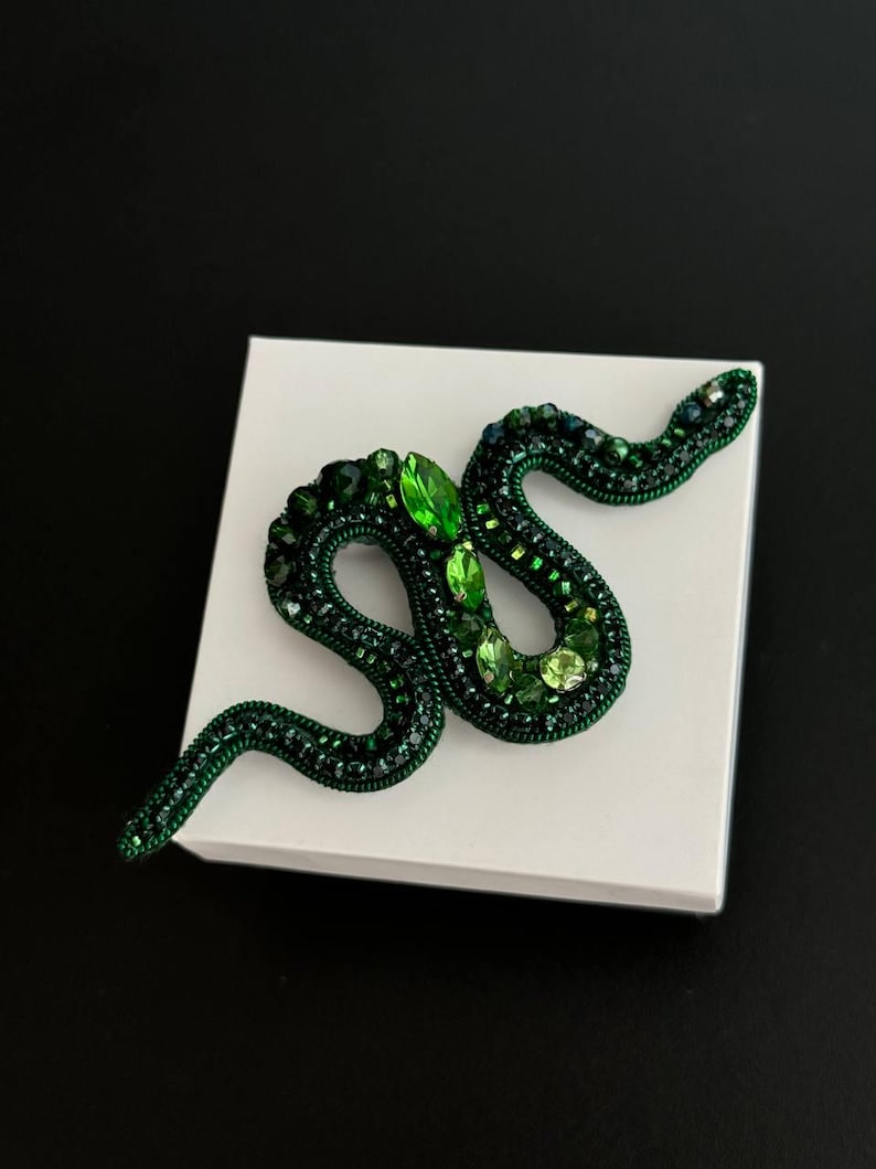 May include: A green snake brooch made of beads and rhinestones. The snake is curled up in a coil shape.